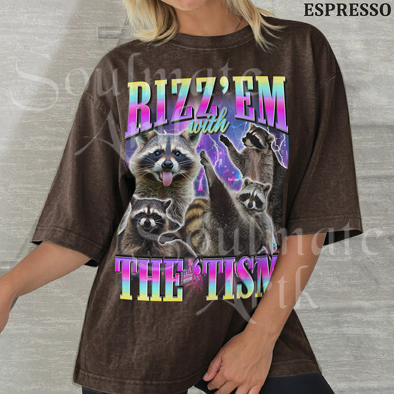 Graphic collage of multiple raccoons over a neon city with bold rainbow text reading RIZZ 'EM WITH THE 'TISM on a washed espresso shirt.