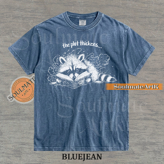 Hand-drawn black and white illustration of a raccoon reading a book with the text The Plot Thickens on a vintage t-shirt