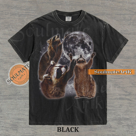 Graphic featuring three detailed raccoons reaching for a giant full moon in a starry space setting on a washed black t-shirt.