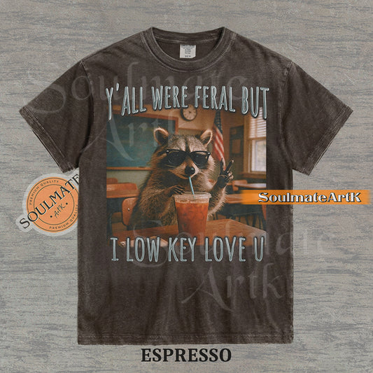 Photorealistic graphic of a raccoon teacher in sunglasses with iced coffee in a classroom under text reading "Y'all Were Feral But I Low Key Love U" on a washed espresso shirt.