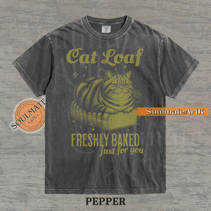 Illustration of a tabby cat sitting on a loaf of bread with text reading "Cat Loaf Freshly Baked just for you" on a washed pepper t-shirt.