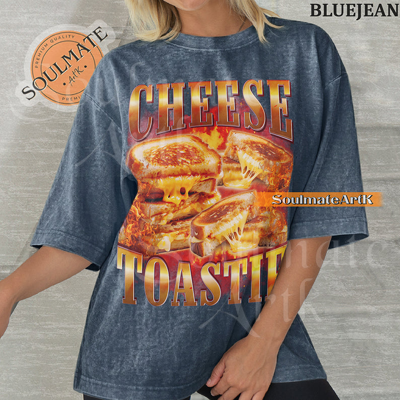 Graphic collage of melting grilled cheese sandwiches surrounded by flames with bold metallic text reading CHEESE TOASTIE on a washed bluejean shirt.