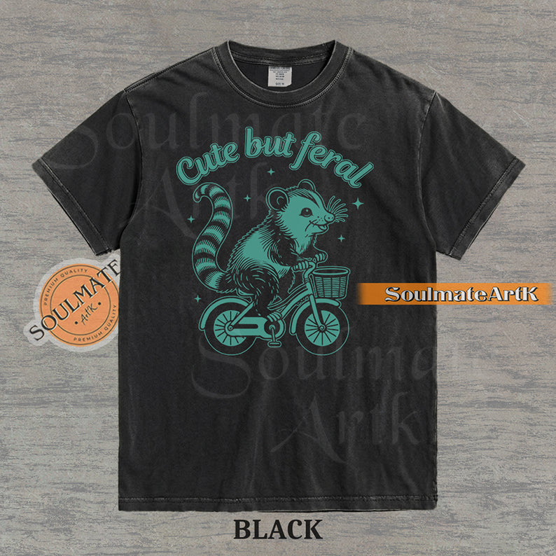 Teal hand-drawn illustration of a cute opossum riding a vintage bicycle with a basket, featuring the text Cute but feral on a washed graphic t-shirt.
