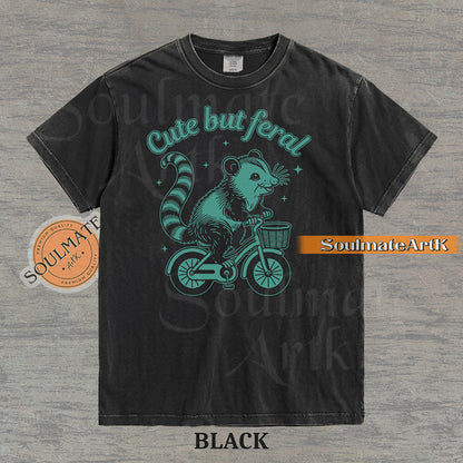 Teal hand-drawn illustration of a cute opossum riding a vintage bicycle with a basket, featuring the text Cute but feral on a washed graphic t-shirt.