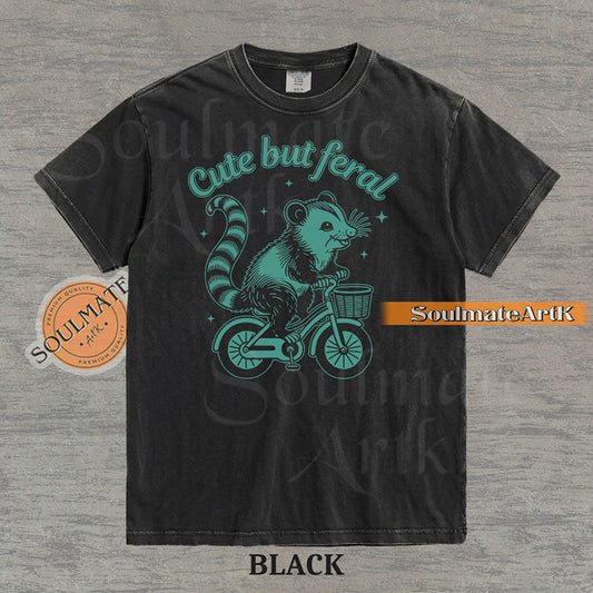 Teal hand-drawn illustration of a cute opossum riding a vintage bicycle with a basket, featuring the text Cute but feral on a washed graphic t-shirt.