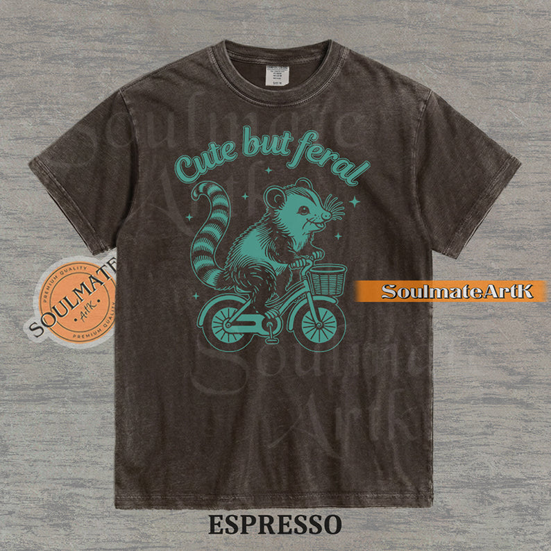 Teal hand-drawn illustration of a cute opossum riding a vintage bicycle with a basket, featuring the text Cute but feral on a washed graphic t-shirt.