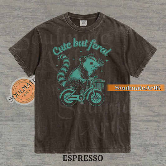 Teal hand-drawn illustration of a cute opossum riding a vintage bicycle with a basket, featuring the text Cute but feral on a washed graphic t-shirt.
