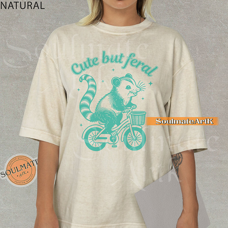 Teal hand-drawn illustration of a cute opossum riding a vintage bicycle with a basket, featuring the text Cute but feral on a washed graphic t-shirt.