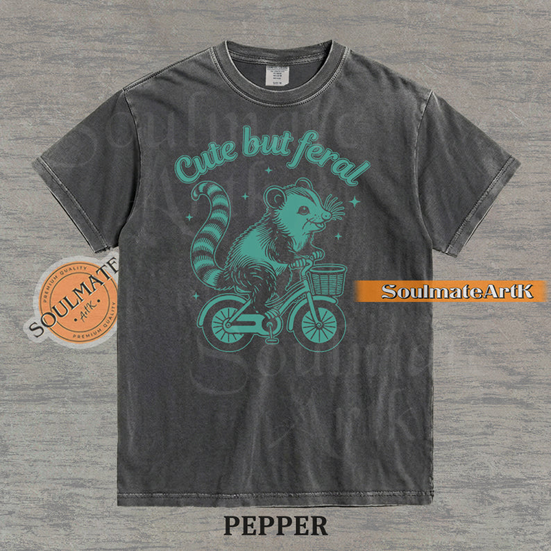 Teal hand-drawn illustration of a cute opossum riding a vintage bicycle with a basket, featuring the text Cute but feral on a washed graphic t-shirt.