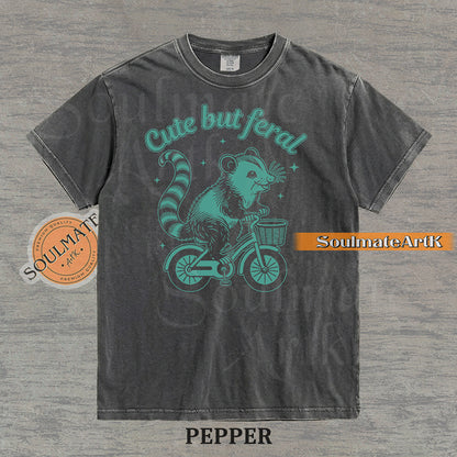 Teal hand-drawn illustration of a cute opossum riding a vintage bicycle with a basket, featuring the text Cute but feral on a washed graphic t-shirt.
