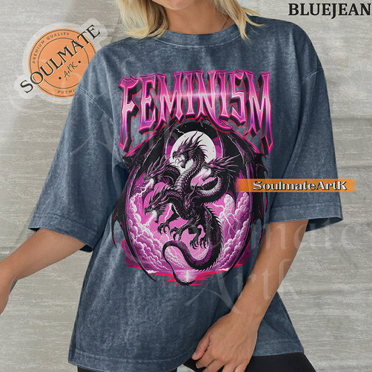Illustration of a multi-headed black and purple dragon in front of a pink moon with bold purple text reading Feminism on a blue-jean colored shirt.