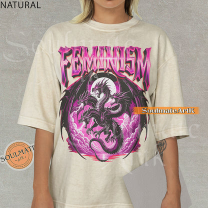 Illustration of a multi-headed black and purple dragon in front of a pink moon with bold purple text reading Feminism on a natural colored shirt.