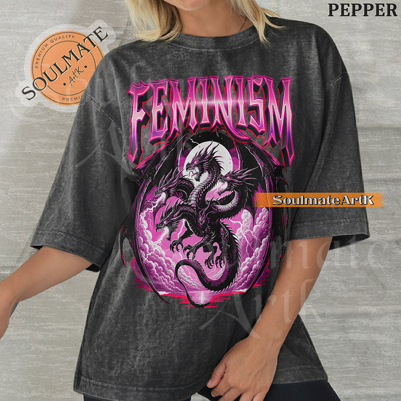 Illustration of a multi-headed black and purple dragon in front of a pink moon with bold purple text reading Feminism on a pepper colored shirt.