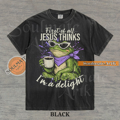 Illustration of a green frog in sunglasses and a purple scarf holding a coffee mug with the text "First of all, Jesus thinks I'm a delight" on a washed black shirt.