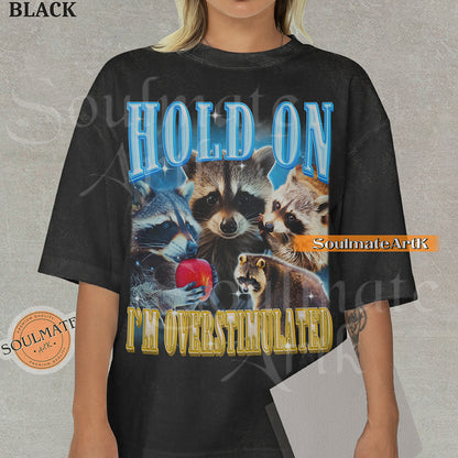 Graphic collage of multiple raccoons with bold blue and gold text reading HOLD ON I'M OVERSTIMULATED on a washed black shirt.