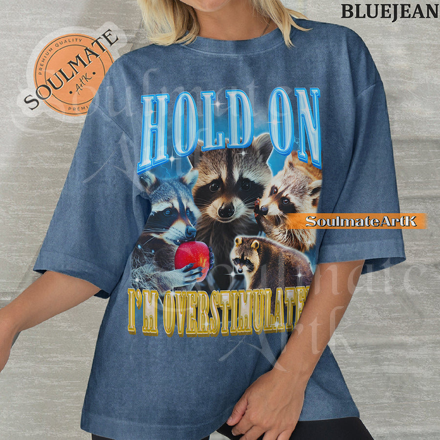 Graphic collage of multiple raccoons with bold blue and gold text reading HOLD ON I'M OVERSTIMULATED on a washed bluejean shirt.