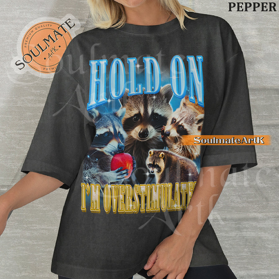 Graphic collage of multiple raccoons with bold blue and gold text reading HOLD ON I'M OVERSTIMULATED on a washed pepper shirt.