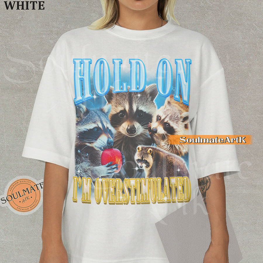 Graphic collage of multiple raccoons with bold blue and gold text reading HOLD ON I'M OVERSTIMULATED on a white shirt.