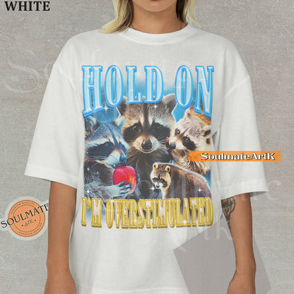 Graphic collage of multiple raccoons with bold blue and gold text reading HOLD ON I'M OVERSTIMULATED on a white shirt.