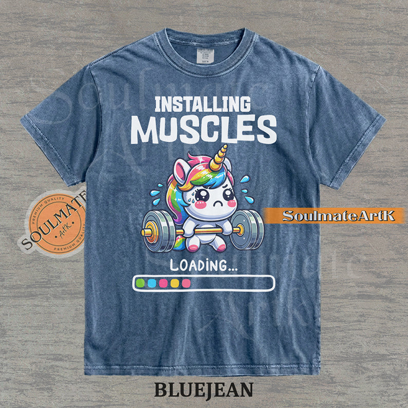 Cute cartoon unicorn lifting a heavy barbell with a loading bar and text reading Installing Muscles on a washed bluejean shirt.