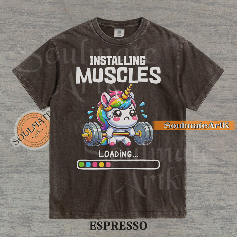 Cute cartoon unicorn lifting a heavy barbell with a loading bar and text reading Installing Muscles on a washed espresso shirt.