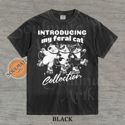 Hand-drawn illustration of three tuxedo cats on roller skates with the text "Introducing my feral cat Collection" on a washed black shirt.