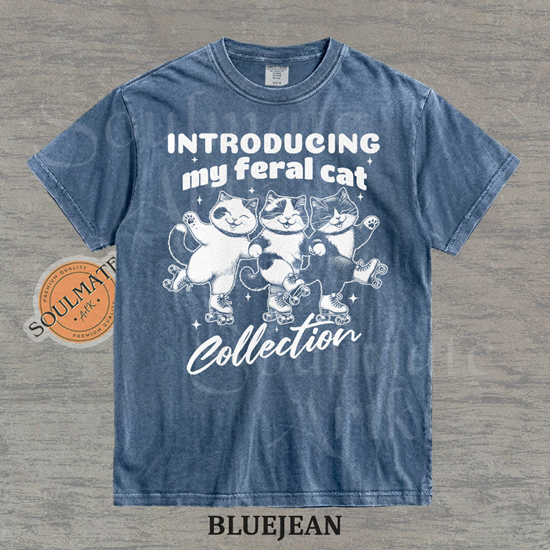 Hand-drawn illustration of three tuxedo cats on roller skates with the text "Introducing my feral cat Collection" on a washed bluejean shirt.