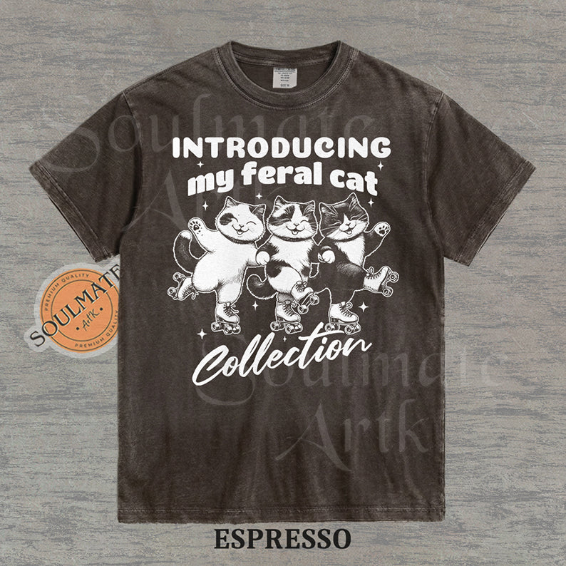 Hand-drawn illustration of three tuxedo cats on roller skates with the text "Introducing my feral cat Collection" on a washed espresso shirt.