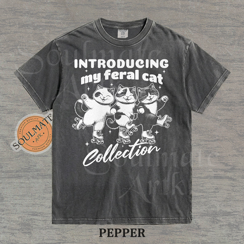 Hand-drawn illustration of three tuxedo cats on roller skates with the text "Introducing my feral cat Collection" on a washed pepper shirt.