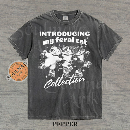 Hand-drawn illustration of three tuxedo cats on roller skates with the text "Introducing my feral cat Collection" on a washed pepper shirt.