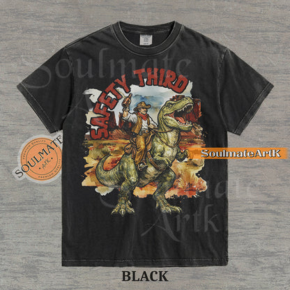 Vintage-style graphic of a cowboy with a hat and drink riding a T-Rex through a desert landscape with red text reading Safety Third on a washed black t-shirt.