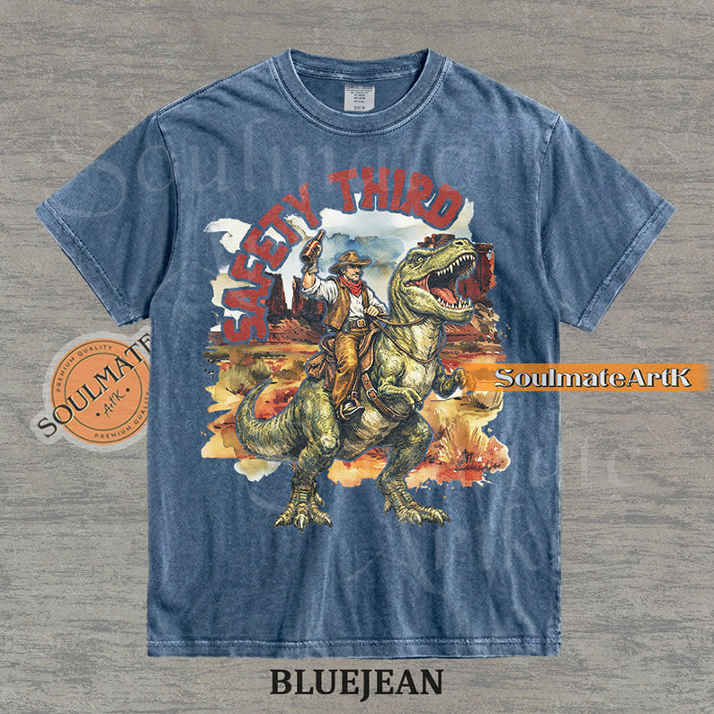 Vintage-style graphic of a cowboy with a hat and drink riding a T-Rex through a desert landscape with red text reading Safety Third on a washed bluejean t-shirt.