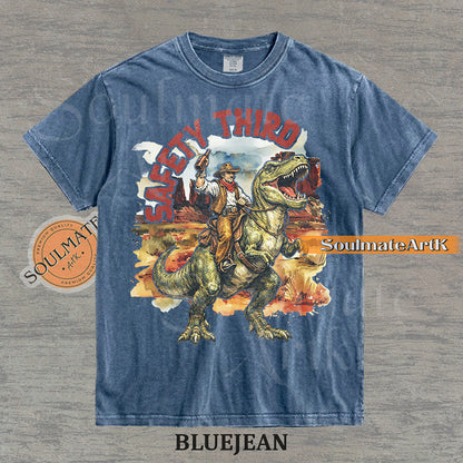 Vintage-style graphic of a cowboy with a hat and drink riding a T-Rex through a desert landscape with red text reading Safety Third on a washed bluejean t-shirt.