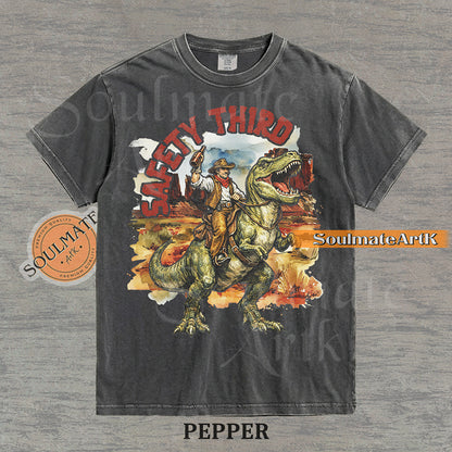Vintage-style graphic of a cowboy with a hat and drink riding a T-Rex through a desert landscape with red text reading Safety Third on a washed pepper t-shirt.