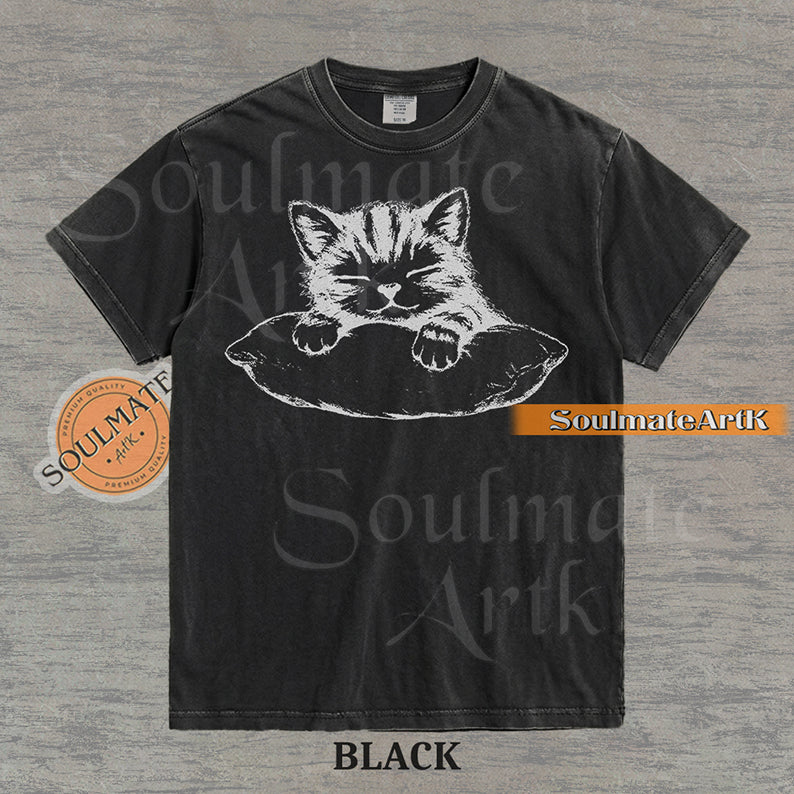 Hand-drawn sketch of a striped kitten sleeping on a pillow in black ink on a black vintage-wash t-shirt.