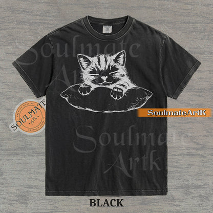 Hand-drawn sketch of a striped kitten sleeping on a pillow in black ink on a black vintage-wash t-shirt.