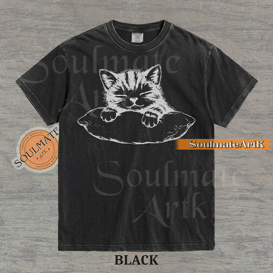 Hand-drawn sketch of a striped kitten sleeping on a pillow in black ink on a black vintage-wash t-shirt.