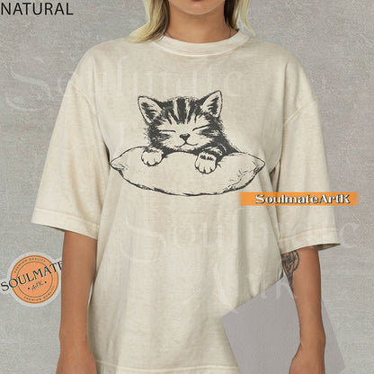 Hand-drawn sketch of a striped kitten sleeping on a pillow in black ink on a natural-colored vintage-wash t-shirt.