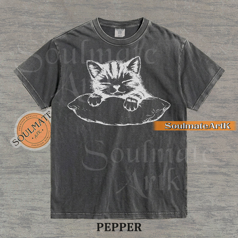 Hand-drawn sketch of a striped kitten sleeping on a pillow in black ink on a pepper vintage-wash t-shirt.