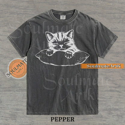 Hand-drawn sketch of a striped kitten sleeping on a pillow in black ink on a pepper vintage-wash t-shirt.