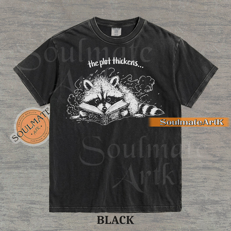 Hand-drawn black and white illustration of a raccoon reading a book with the text The Plot Thickens on a vintage t-shirt