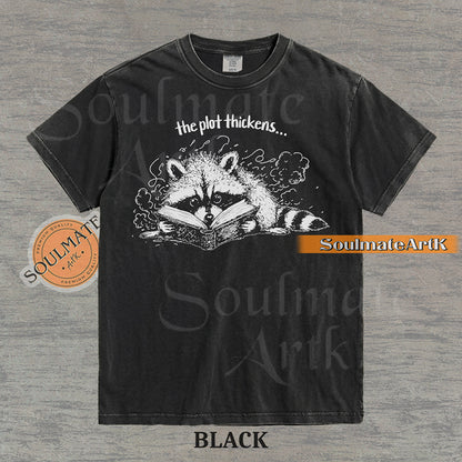 Hand-drawn black and white illustration of a raccoon reading a book with the text The Plot Thickens on a vintage t-shirt