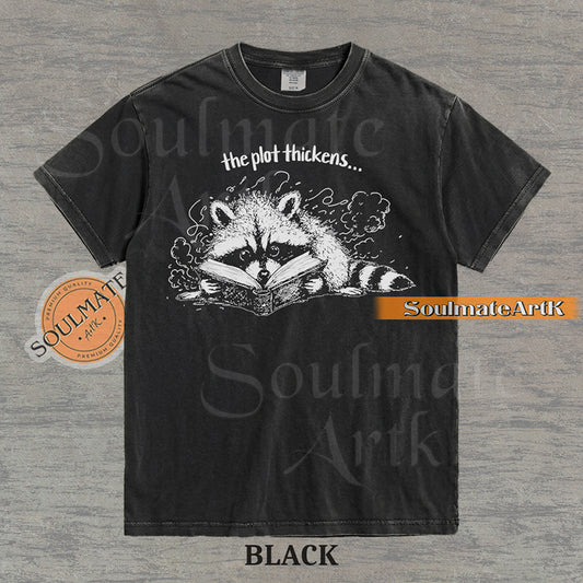 Hand-drawn black and white illustration of a raccoon reading a book with the text The Plot Thickens on a vintage t-shirt
