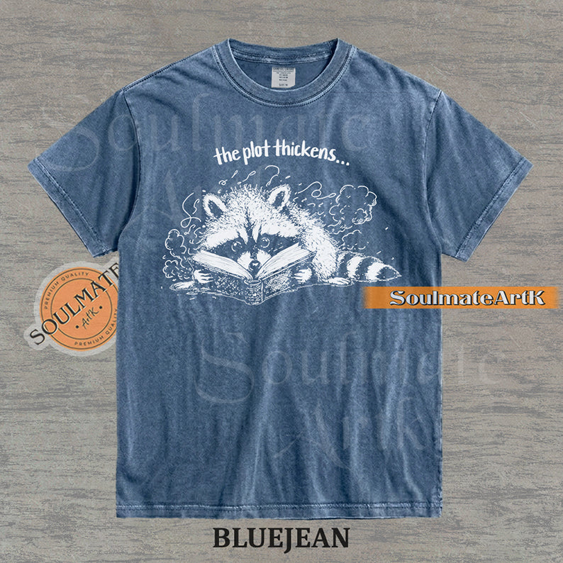 Hand-drawn black and white illustration of a raccoon reading a book with the text The Plot Thickens on a vintage t-shirt