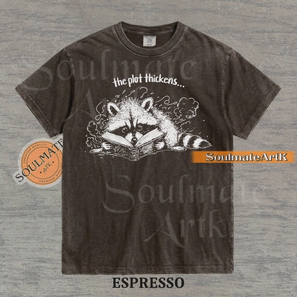 Hand-drawn black and white illustration of a raccoon reading a book with the text The Plot Thickens on a vintage t-shirt