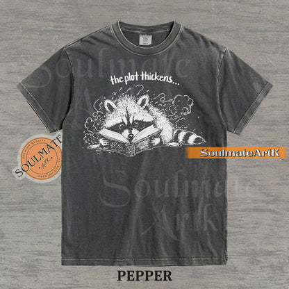 Hand-drawn black and white illustration of a raccoon reading a book with the text The Plot Thickens on a vintage t-shirt