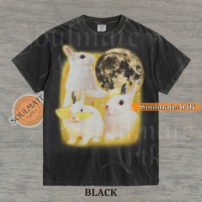 Graphic of three white rabbits—one wearing yellow sunglasses—next to a full moon in a golden space setting on a washed black t-shirt.