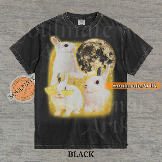 Graphic of three white rabbits—one wearing yellow sunglasses—next to a full moon in a golden space setting on a washed black t-shirt.