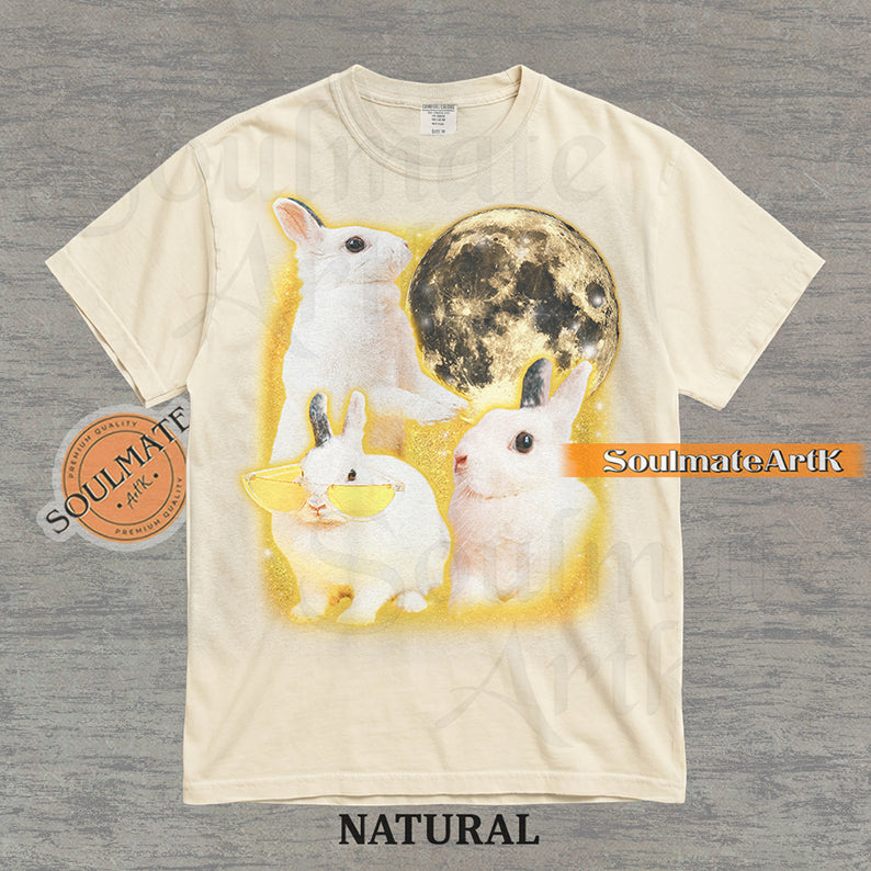 Graphic of three white rabbits—one wearing yellow sunglasses—next to a full moon in a golden space setting on a natural t-shirt.