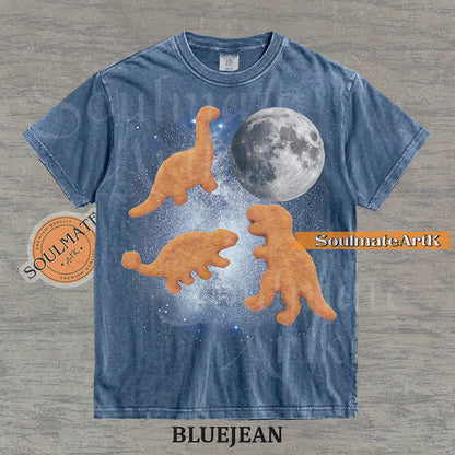 Vintage-style graphic of three golden dinosaur nuggets floating in a blue space nebula with a large moon on a washed bluejean t-shirt.